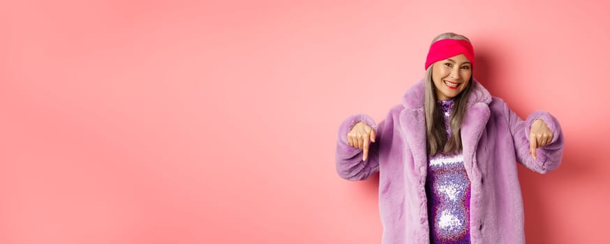 Fashion and shopping concept. Smiling senior asian woman pointing fingers down, showing store advertisement on bottom, wearing stylish purple winter coat, pink background.