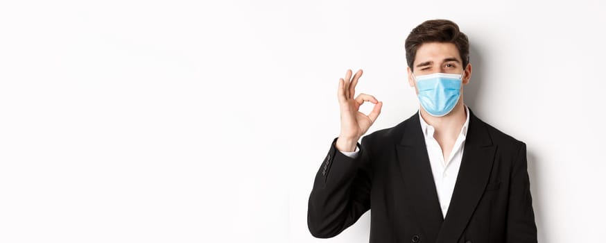 Concept of covid-19, business and social distancing. Close-up of good-looking businessman in trendy suit and medical mask, showing okay sign and winking, standing against white background.