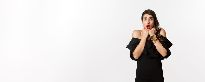 Portrait of glamour woman looking scared and shocked at camera, staring at something with fear, standing in black dress against white background