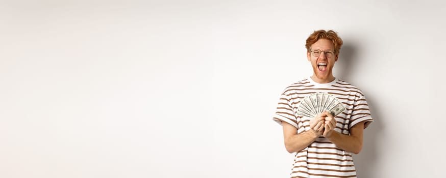 Lucky young man with red hair showing dollars, winning money and screaming of happiness, holding prize cash, standing over white background