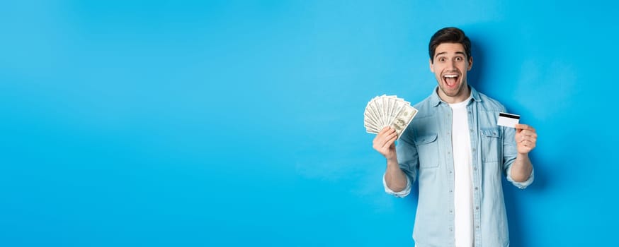 Happy attractive man looking amazed, showing cash and credit card, concept of banks, credit and finance. Blue studio background