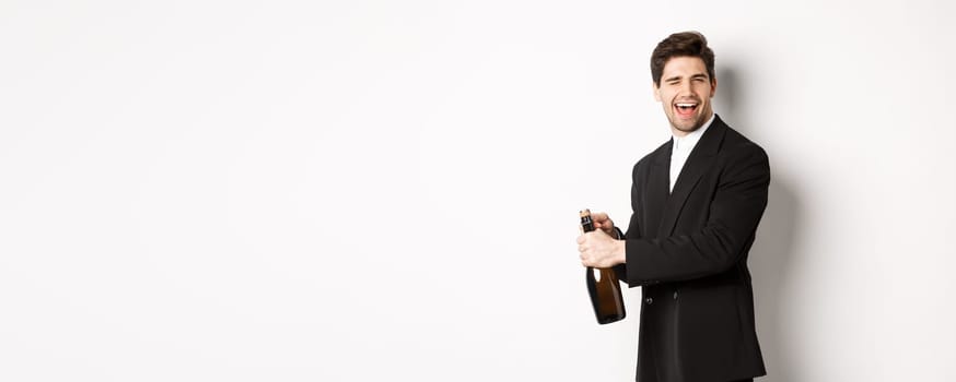 Portrait of attractive man in black suit, winking at camera and opening bottle of champagne, celebrating new year, standing against white background