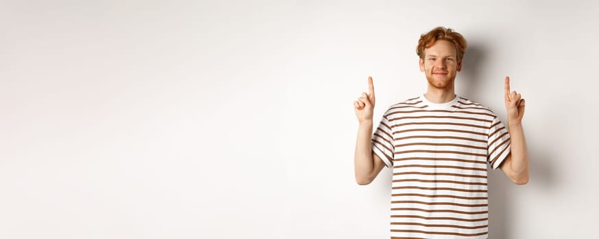 Smiling redhead guy with beard, pointing fingers up and showing advertisement, standing over white background