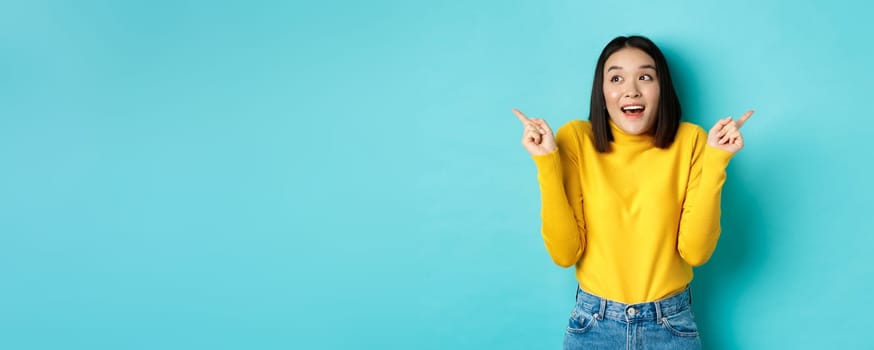 Beauty and fashion concept. Excited asian girl in yellow pullover making choice, pointing fingers sideways and choosing between two options, standing against blue background