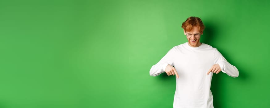 Handsome redhead man in glasses and white long-sleeve checking out advertisement, pointing fingers and looking down with pleased smile, standing over green background