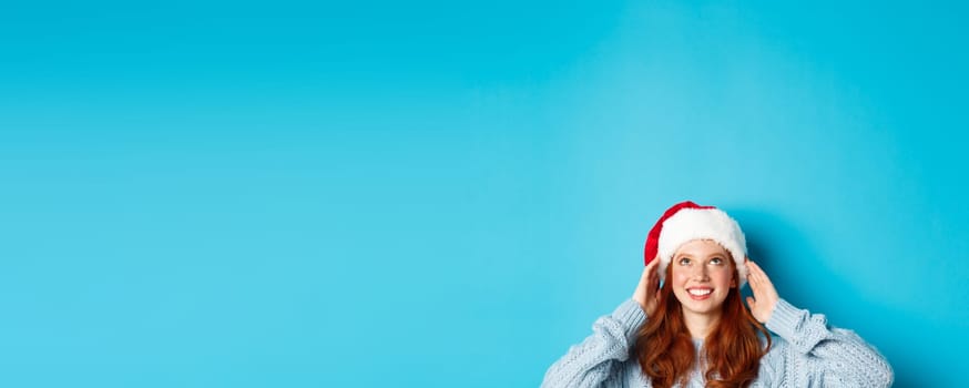 Winter holidays and Christmas eve concept. Head of cute redhead girl in santa hat, appear from bottom and looking up at copy space, staring logo, standing over blue background
