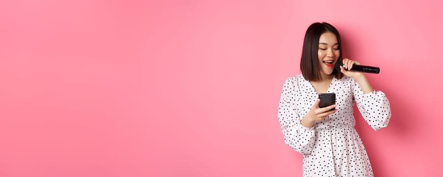 Cute asian woman reading lyrics in smartphone, singing with microphone, standing in trendy dress over pink background