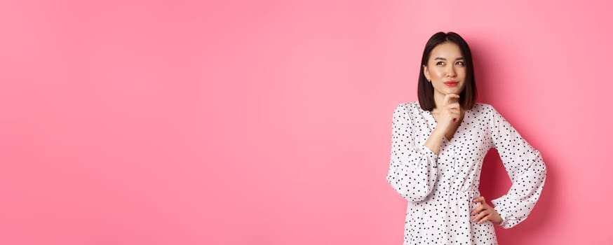 Thoughtful happy asian woman making decision, smiling satisfied and looking at upper left logo, thinking or choosing, standing in dress against pink background