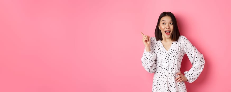 Excited asian woman in stylish dress, gasping amused, pointing finger upper left corner, showing interesting promo offer, standing over pink background