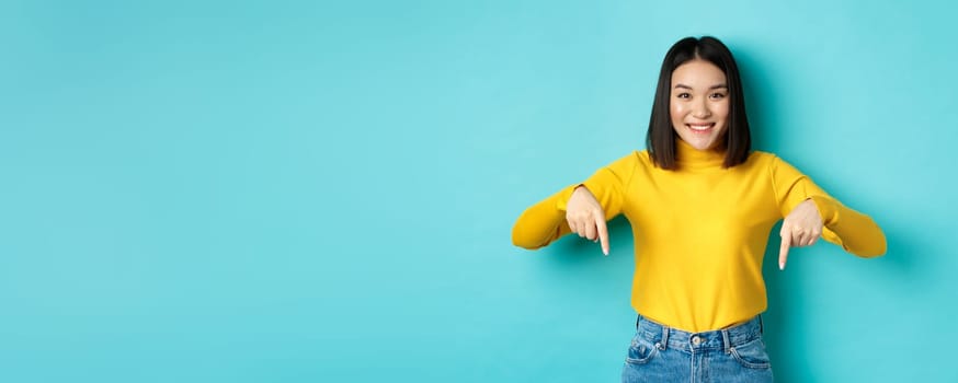 Shopping concept. Beautiful korean girl with happy smile, pointing fingers down at banner, standing against blue background