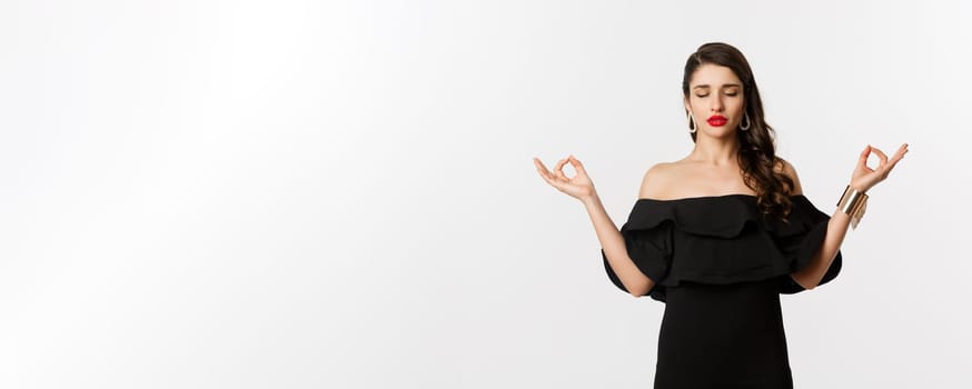 Calm and patient young brunette female meditating, standing in black dress with hands in zen gesture, standing against white background