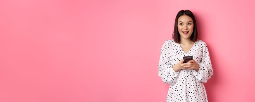 Cute asian woman thinking and smiling, looking left dreamy while messaging on smartphone, browing online stores, standing over pink background
