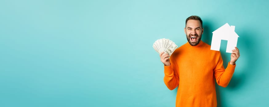 Real estate concept and mortgage concept. Ecstatic caucasian man holding money dollars and house maket, shouting for joy, standing over blue background