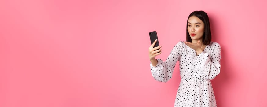 Beautiful asian girl using photo filters app and taking selfie on smartphone, posing in cute dress against pink background