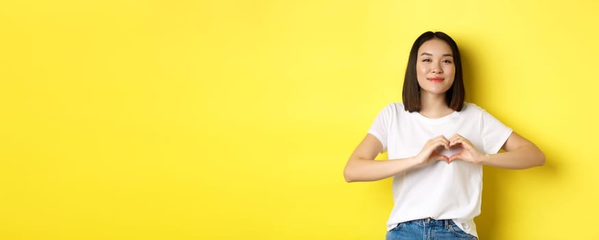 Beautiful asian woman showing I love you heart gesture, smiling at camera, standing against yellow background. Concept of valentines day and romance