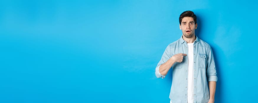 Nervous man pointing at himself and looking confused, standing in casual clothes over blue background