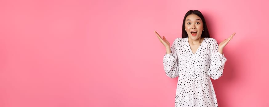 Happy and surprised asian woman rejoicing, spread hands and gasping amazed, looking with excitement and disbelief, standing over pink background