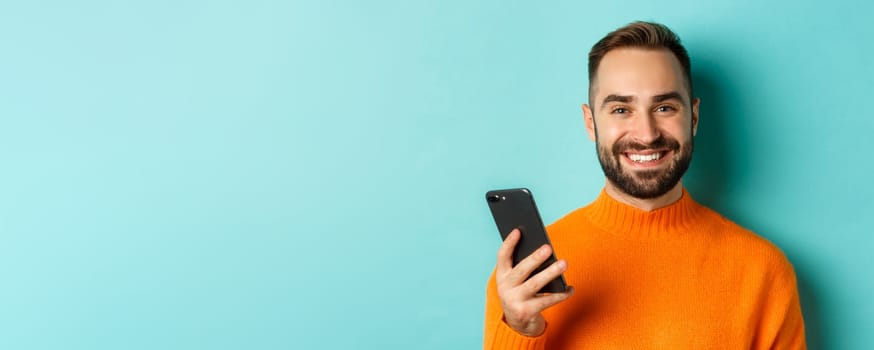 Close-up of happy handsome man writing message on mobile phone, holding smartphone and smiling, standing against turquoise background