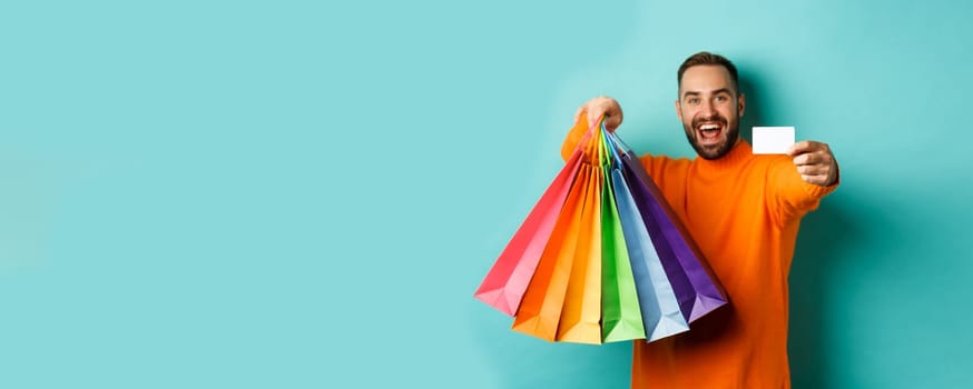 Happy aduly man showing credit card and shopping bags, standing against turquoise background