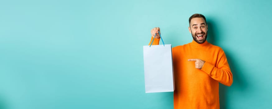 Handsome smiling man pointing finger at shopping bag, buying in stores, standing satisfied over blue background