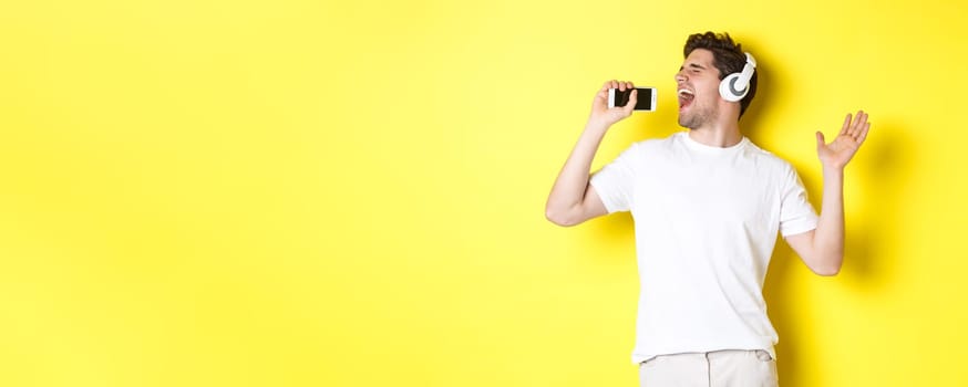 Happy guy playing karaoke app in headphones, singing into smartphone microphone, standing over yellow background