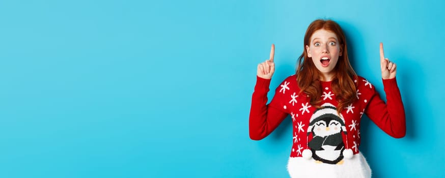 Winter holidays and celebration concept. Amazed redhead girl showing advertisement, pointing at christmas logo, standing excited over blue background.