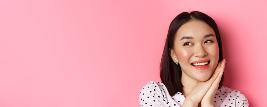 Beauty and skin care concept. Headshot of adorable and dreamy asian woman looking left, smiling and imaging, standing against pink background