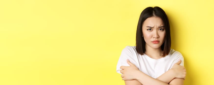 Sad timid girl comforting herself, hugging body and frowning upset, standing again yellow background