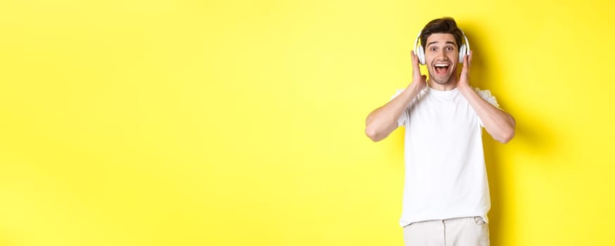 Man in headphones looking surprised and happy, listening awesome song, standing over yellow background