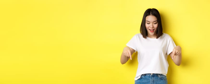 Beauty and fashion concept. Beautiful asian woman in white t-shirt pointing fingers down, demonstrate logo standing over yellow background
