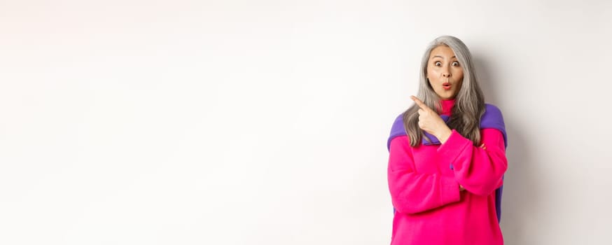 Portrait of impressed asian lady checking out promo offer, pointing at upper left corner and staring at camera amazed, standing in trendy pink sweater over white background