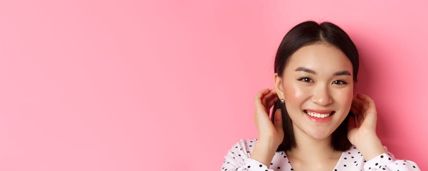 Beauty and skin care concept. Close-up of adorable smiling asian woman tuck hair behind ears, blushing and gazing at camera, standing over pink background