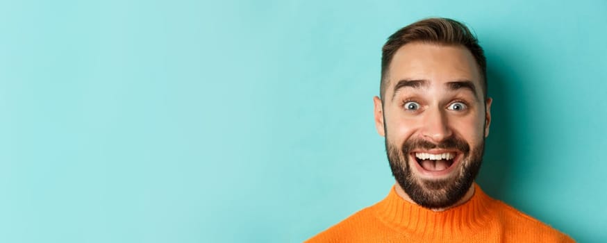 Headshot of handsome caucasian man with beard smiling happy at camera, staring amazed, standing in orange sweater against turquoise background