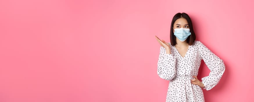 Covid-19, quarantine and lifestyle concept. Lovely asian woman in face mask raising hand, standing in dress over pink background