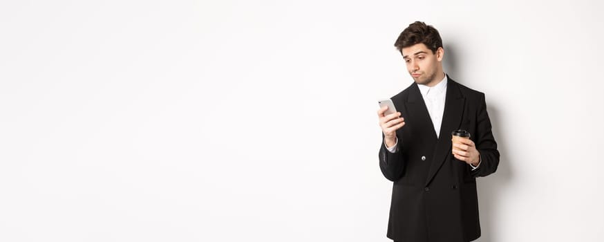 Portrait of thoughtful handsome businessman, drinking coffee and browsing in internet, looking at smartphone screen, standing against white background