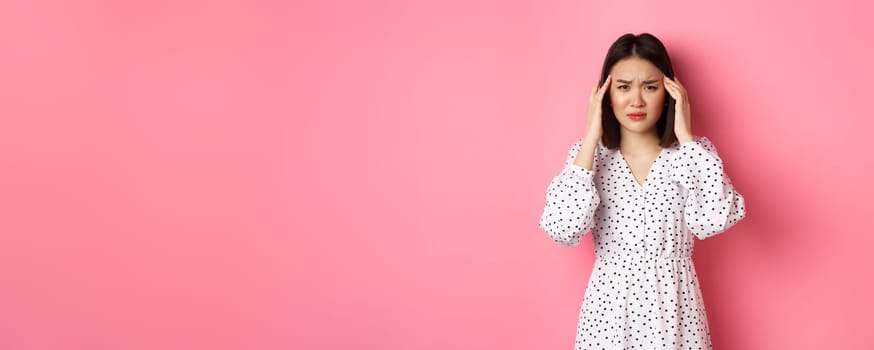 Image of upset asian woman having headache, feeling unwell or dizzy, close eyes and massaging head, suffering migraine, standing over pink background