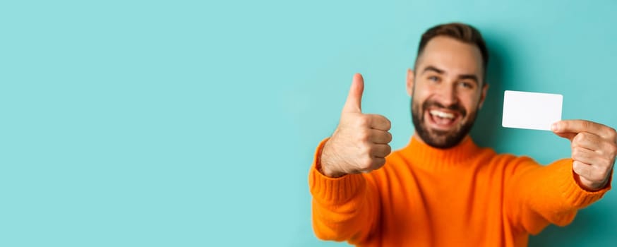 Close-up of handsome caucasian man going on shopping, showing credit card and thumb up in approval, standing over turquoise background