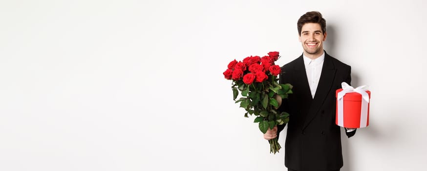 Concept of holidays, relationship and celebration. Handsome boyfriend in black suit, holding bouquet of red roses and a gift, wishing merry christmas, standing over white background