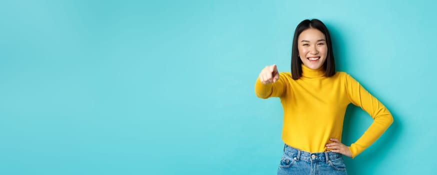 Beauty and fashion concept. Cheerful asian woman laughing and smiling, pointing finger at camera, choose you, standing over blue background.