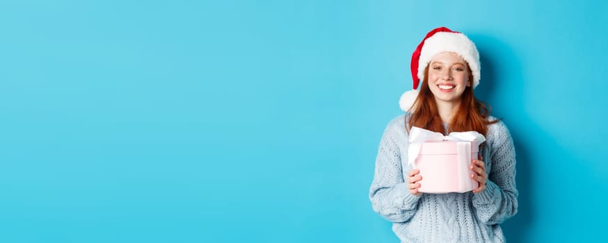 Winter holidays and Christmas Eve concept. Smiling redhead girl in sweater and Santa hat, holding New Year gift and looking at camera, standing against blue background