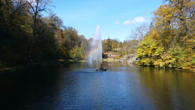 Fountain with rainbow in middle of lake between trees with yellow leaves in park