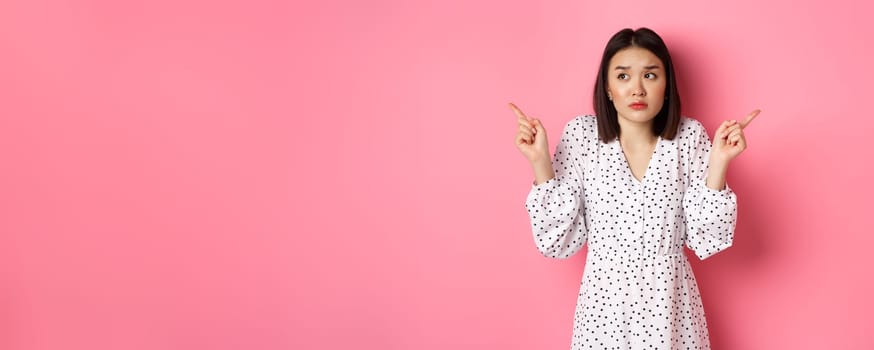 Confused asian woman hesitating, shrugging and pointing hands two sides, staring indecisive and need help with choice, standing over pink background