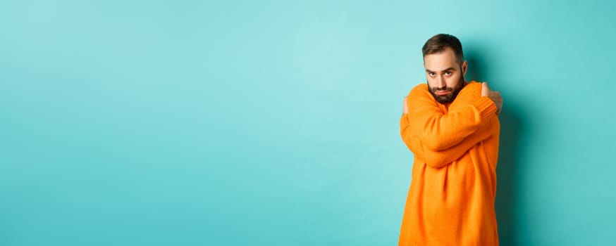 Timid man feeling offended and defensive, hugging himself and looking suspicious at camera, standing over light blue background