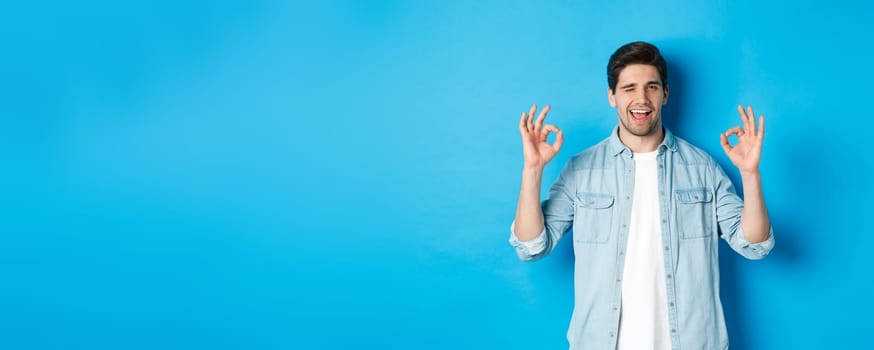Relaxed and confident man showing ok signs and winking, everything okay gesture, standing against blue background