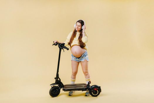 Pregnant girl in yellow clothes and headphones on an electric scooter on an isolated yellow background