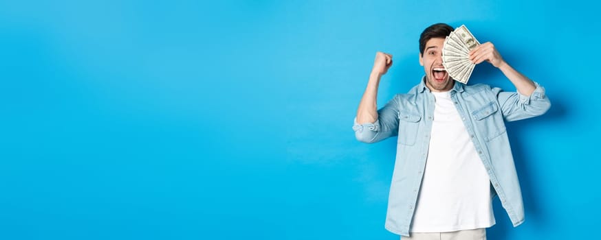 Successful young man earn money, making fist pump and showing cash, winning prize or receive credit, standing over blue background