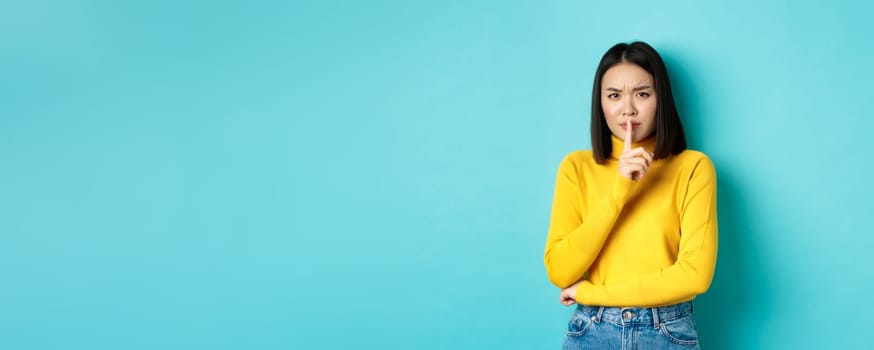 Disappointed asian woman telling to be quiet, scolding loud person with hush gesture, shushing at camera and frowning upset, standing over blue background