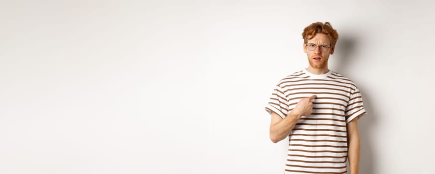 Confused redhead man in glasses pointing at himself, looking questioned at camera, white background