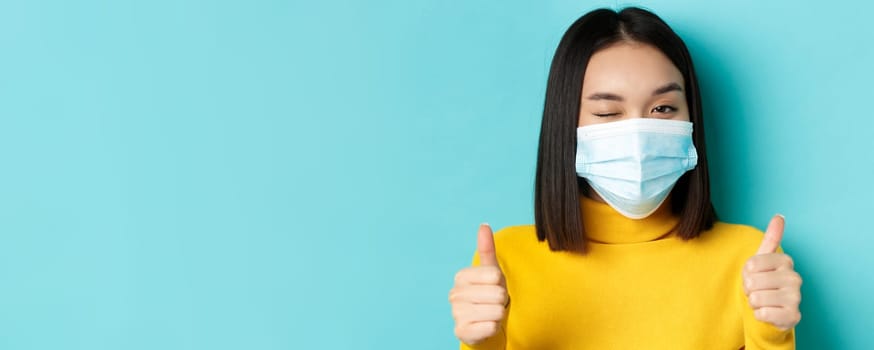 Covid-19, social distancing and pandemic concept. Cute asian girl in medical mask winking at camera, showing thumbs up, good job gesture, praise nice work, blue background