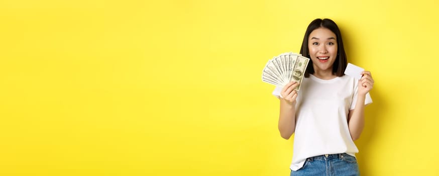 Beautiful asian woman with short dark hair, wearing white t-shirt, showing money in dollars and plastic credit card, standing over yellow background